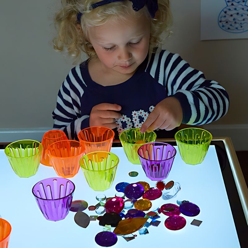 TTS translucent stacking cups glowing in vibrant colours on an LED light table, ideal for early years visual sensory exploration and fine motor development