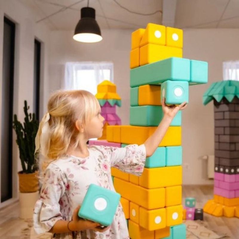 Girl picking up a large 10cm TUKI BIG magnetic foam block from the Builder 150 set, showing the whole-body proprioceptive engagement and bilateral coordination that large-format floor building with the 150-piece largest BUILDER set develops
