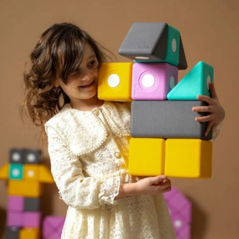 Girl lifting a large 10cm TUKI BIG magnetic foam block from the Builder 26 set, demonstrating the bilateral coordination, proprioceptive loading, and whole-body gross motor engagement that large-format floor building with the 26-piece BIG set develops