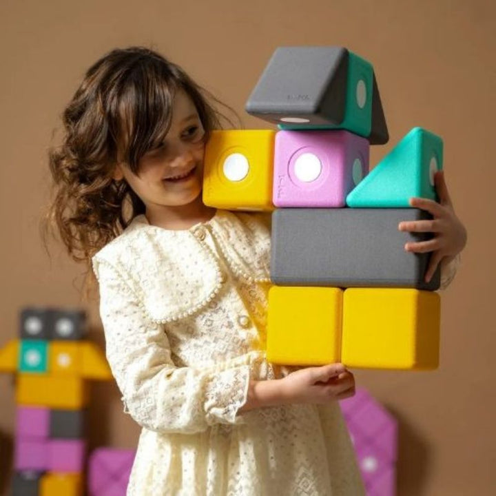 Girl lifting a large 10cm TUKI BIG magnetic foam block from the Builder 76 set, showing the bilateral coordination, proprioceptive loading, and whole-body motor development that large-format floor building with the 76-piece EN71-certified foam blocks develops