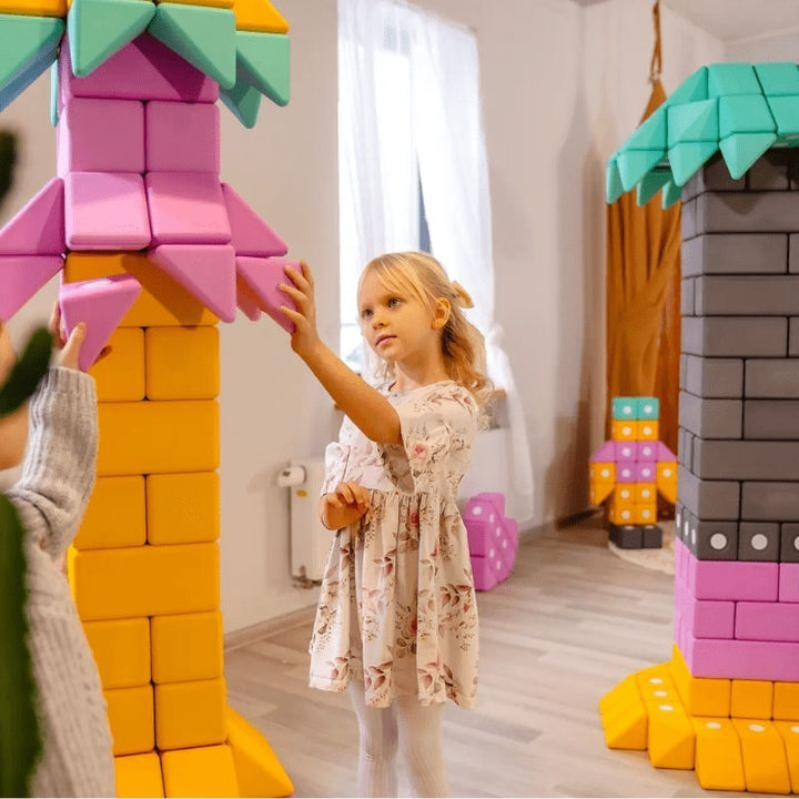 Young girl picking up a large foam block from the TUKI Smart 240 set, demonstrating the lightweight design and gross motor engagement during sensory building play