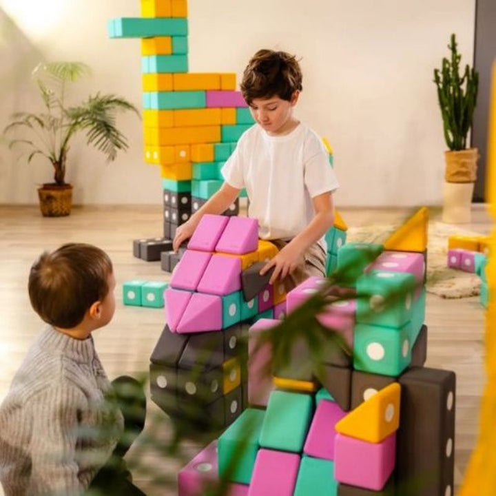 Boys sitting on and riding a structure built from the TUKI BIG Smart Magnetic Blocks Smart 160, demonstrating the physical inhabitation scale that 160 large-format 10cm foam blocks produces - structures large enough to support a child's weight and function as play equipment