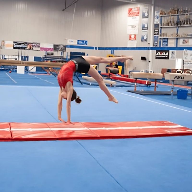 Gymnast performing a cartwheel on the Tumbl Trak Hopscotch Beam Mat beam stripe side in a competitive gymnastics club, demonstrating straight-line tumbling and balance beam skill training on the reversible gymnastics floor mat