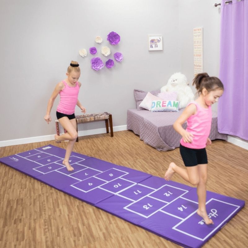 Two children playing hopscotch on the purple side of the Tumbl Trak Hopscotch Beam Mat in a home bedroom, showing the mat's compact 3x12ft footprint for home use and its numbered hopscotch pattern for active movement play