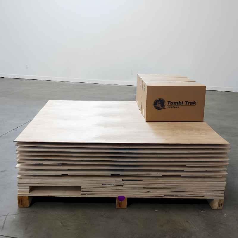 Tumbl Trak spring floor Baltic Birch panels stacked flat on a shipping pallet alongside the included Assembly Care Package box, showing how the semi-assembled system arrives ready for installation