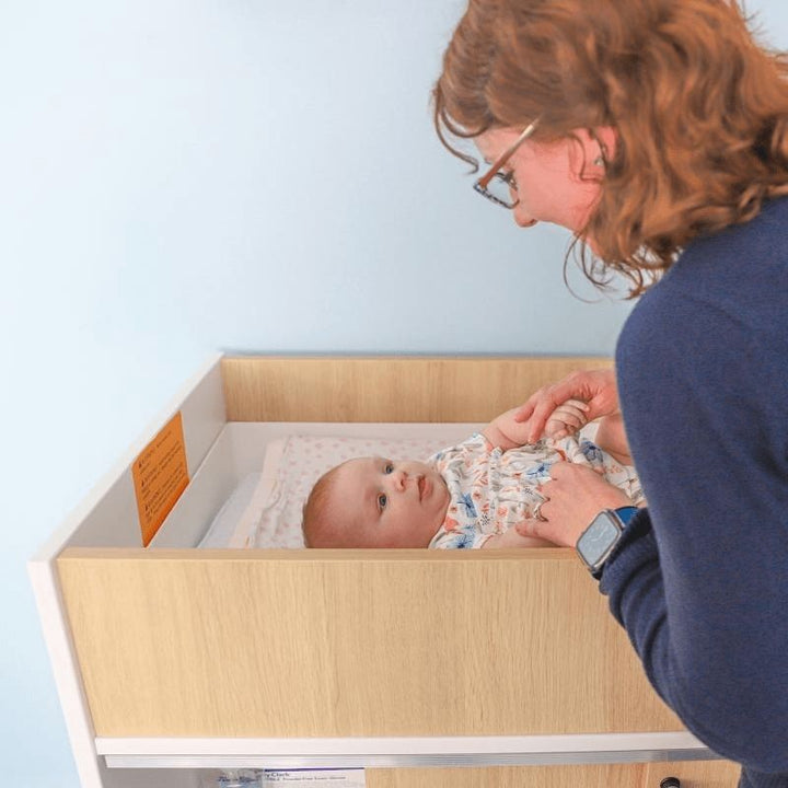 Close-up of mother and baby at the Whitney Brothers elevatED Infant Changing Cabinet, showing the washable changing pad and safety strap included with the antimicrobial changing station
