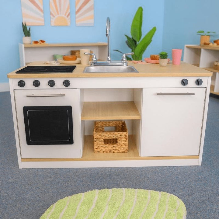 Whitney Brothers elevatED Kitchen Island Breakfast Bar in a classroom or play space showing the full front kitchen face with stovetop, sink, oven, dishwasher, and open centre shelves