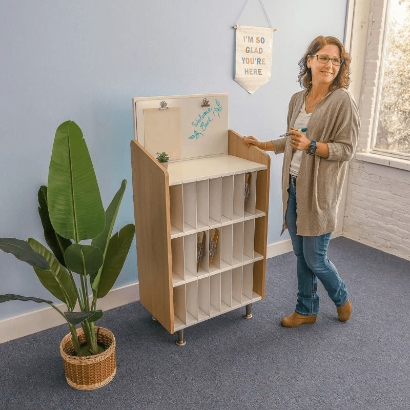 Adult using the Whitney Brothers elevatED Mail and Message Center in a classroom setting, showing the 24 vertical slots, removable dividers, and the dry-erase board with clips at the top