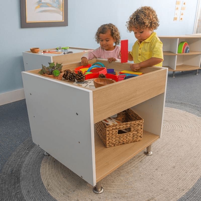 Toddler playing at the Whitney Brothers elevatED Toddler Discovery Table, showing the low 24" first-walker height and the contemporary white and textured white oak two-tone elevatED design