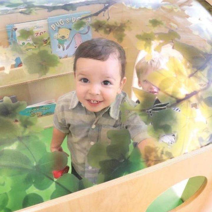 Child reading inside the Whitney Brothers Nature Reading Haven with optional Green Floor Mat, showing the cosy interior with tree canopy overhead and built-in book display shelves