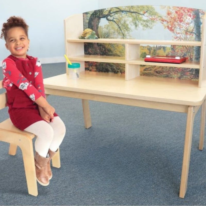 Happy child seated at the Whitney Brothers Nature View Writing Center, showing the forest scene acrylic back panel and the 20" desk surface of the award-winning biophilic writing desk
