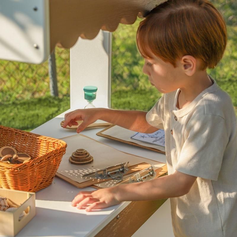 Close-up of child engaged in outdoor pretend play at the Whitney Brothers outdoorED Market Stand counter, showing child-scaled stand height and textured woodgrain maple finish