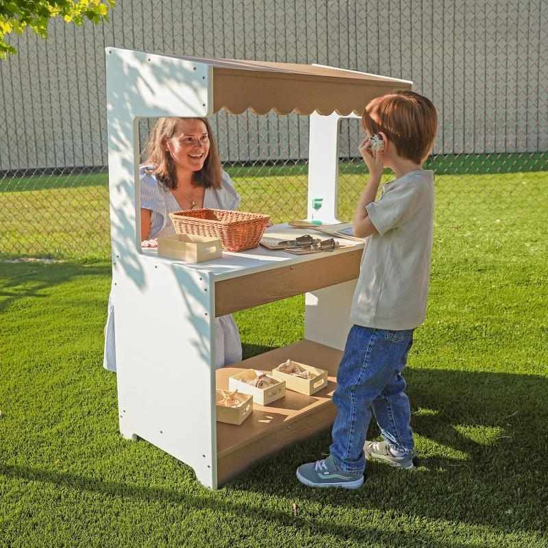 Mother and child engaged in outdoor dramatic play at the Whitney Brothers outdoorED Market Stand, showing sloped awning, writable front panel, and weatherproof HDPE construction