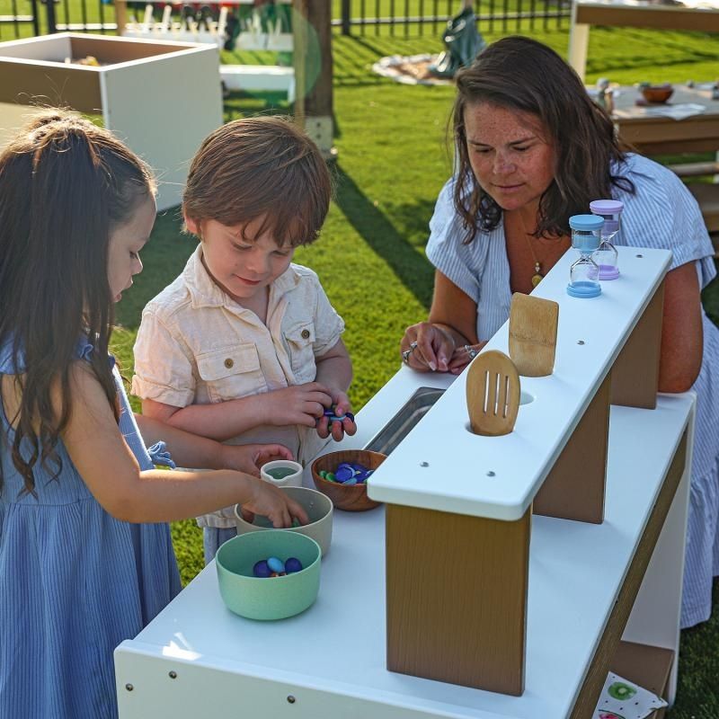 Mother and two children collaborating at the Whitney Brothers outdoorED outdoor mud kitchen, showing multi-child play surface and removable stainless steel sink
