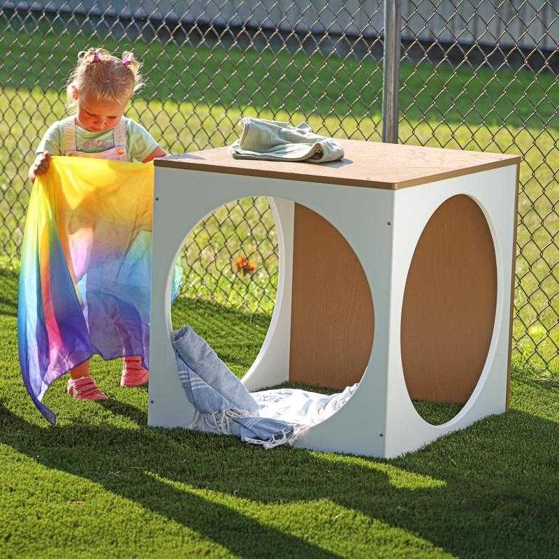 Child engaging in active outdoor play around the Whitney Brothers outdoorED Play House Cube, showing weatherproof HDPE construction and woodgrain maple finish in outdoor preschool setting