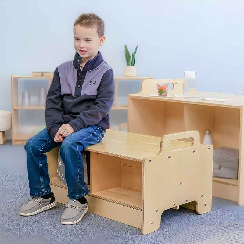 Child seated on the Whitney Brothers Stack-Able Bench in an early learning classroom, showing the natural birch plywood construction and under-seat divided storage compartments